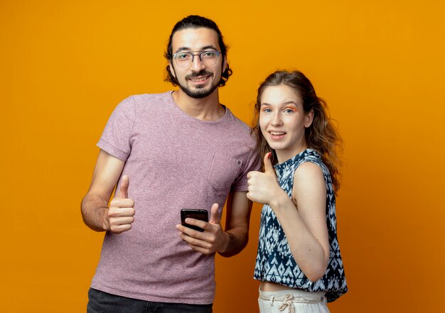 Pareja joven mirando a la cámara sonriendo feliz y positivo mostrando Thumbs up de pie sobre fondo naranja