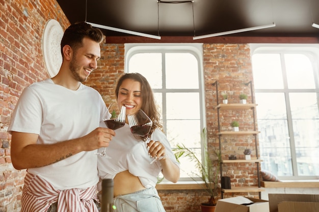 Pareja joven se mudó a una nueva casa o apartamento. Beber vino tinto, acariciar y relajarse después de limpiar y desempacar. Luce feliz y confiado. Familia, mudanza, relaciones, primer concepto de hogar.