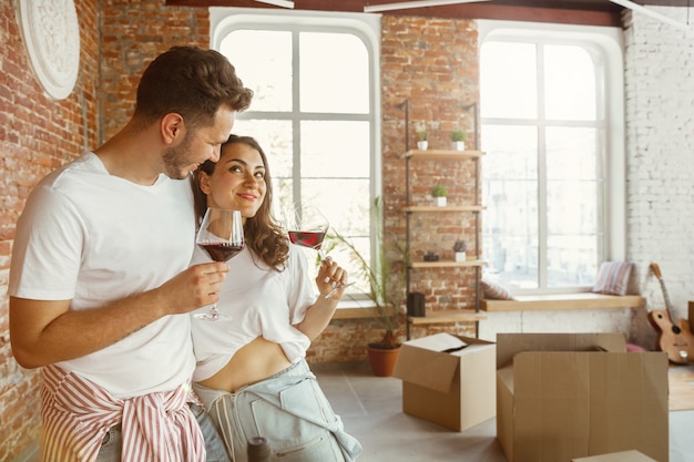 Pareja joven se mudó a una nueva casa o apartamento. Beber vino tinto, acariciar y relajarse después de limpiar y desempacar