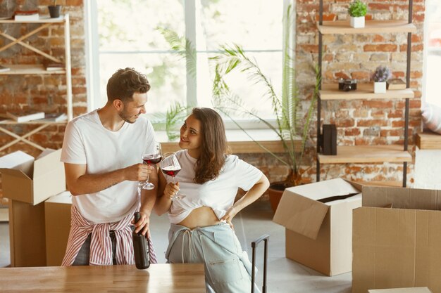 Pareja joven se mudó a una nueva casa o apartamento. Beber vino tinto, sonreír y relajarse después de limpiar y desempacar. Luce feliz y confiado. Familia, mudanza, relaciones, primer concepto de hogar.