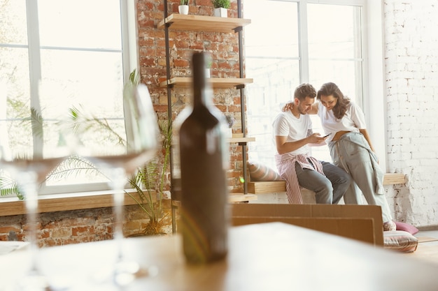 Pareja joven se mudó a una nueva casa o apartamento. Beber vino tinto, usar el teléfono inteligente y relajarse después de limpiar y desempacar. Luce feliz y confiado. Familia, mudanza, concepto de relaciones.