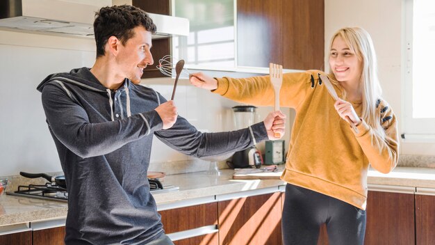 Pareja joven peleando con espátula de madera; cuchara y batidor en la cocina