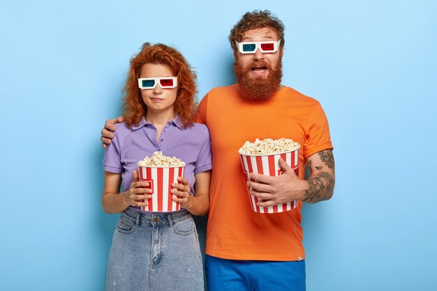 Pareja joven pelirroja pasa tiempo libre en el cine 3D, come palomitas de maíz, usa anteojos especiales para efectos visuales, chico barbudo emocionado abraza a una novia encantadora que tiene expresión aburrida, no le gusta la película