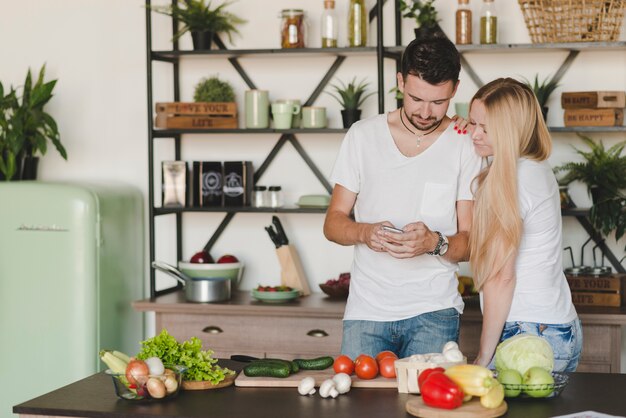 Pareja joven de pie detrás del mostrador de la cocina con teléfono móvil