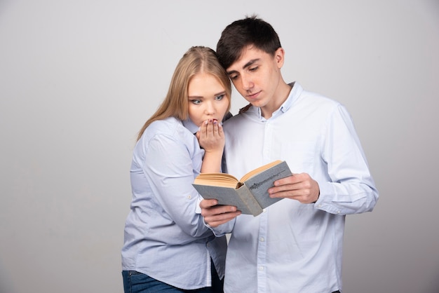 Pareja joven de pie y leyendo un libro sobre la pared gris