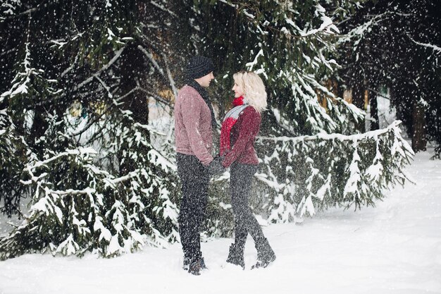 Pareja joven de pie y mirándose en el bosque nevando. Esposa rubia y su esposo con suéteres rojos. Mujer con cabello voluminoso tocando a su pareja. Concepto de ternura.