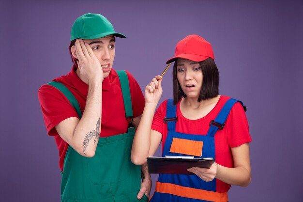 Pareja joven preocupada en uniforme de trabajador de la construcción y gorra chica sosteniendo lápiz y portapapeles tocando la cabeza con lápiz mirando al chico del portapapeles manteniendo la mano en la cara mirando al lado