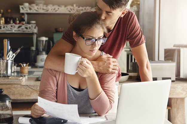 Pareja joven con problema de crédito en el banco. Hombre solidario abrazando y besando a su infeliz esposa en la cabeza mientras ella está sentada en la mesa de la cocina frente a la computadora portátil