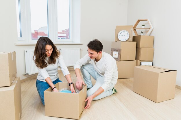 Pareja joven que se muda a un nuevo lugar sentado cerca de una caja de cartón desempacando platos