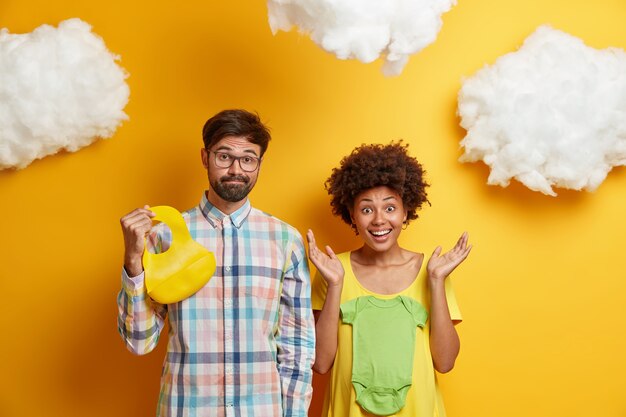 Pareja joven de raza mixta espera bebé, compra ropa para el futuro niño, posa con camiseta y babero de goma, se prepara para dar a luz, aislado sobre amarillo. Felices futuros padres posan en casa.