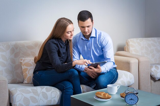 Pareja joven riéndose, sentada en un sofá, compartiendo información, mostrada en una tableta electrónica y tomando café