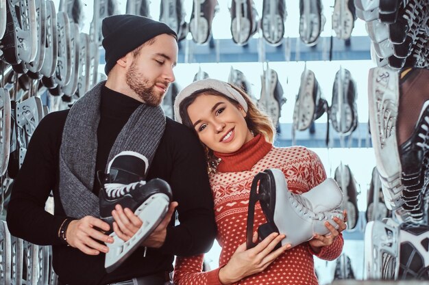 Una pareja joven con ropa abrigada parada cerca del estante con muchos pares de patines, eligiendo su tamaño, preparándose para patinar en la pista de hielo.