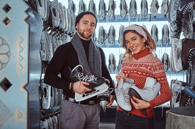 Una pareja joven con ropa abrigada parada cerca del estante con muchos pares de patines, eligiendo su tamaño, preparándose para patinar en la pista de hielo.