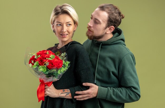 Pareja joven en ropa casual hombre feliz abrazando a su hermosa mujer con flores celebrando el día de San Valentín de pie sobre la pared verde