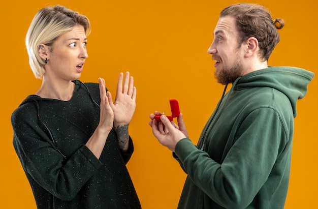 Foto gratuita pareja joven en ropa casual hombre feliz haciendo propuesta con anillo de compromiso en caja roja a su concepto de novia confundida y disgustada de pie sobre la pared naranja