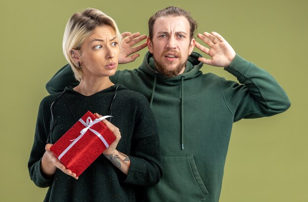 Pareja joven en ropa casual, hombre y mujer con presente mirando confundido y disgustado celebrando el día de San Valentín de pie sobre fondo verde