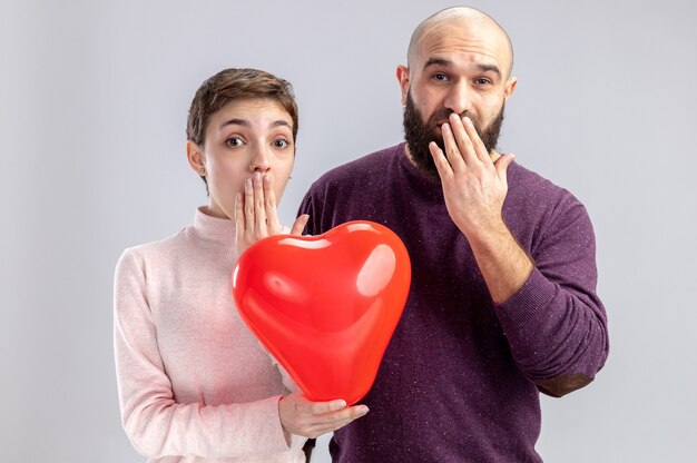 Pareja joven en ropa casual hombre y mujer sosteniendo un globo en forma de corazón mirando a la cámara asombrado y sorprendido cubriendo la boca con las manos celebrando el día de San Valentín de pie sobre fondo blanco.