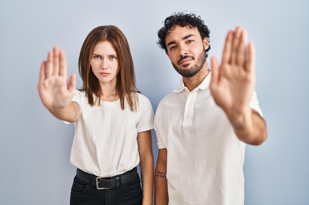 Pareja joven con ropa informal de pie juntos haciendo dejar de cantar con la palma de la mano. expresión de advertencia con gesto negativo y serio en la cara.