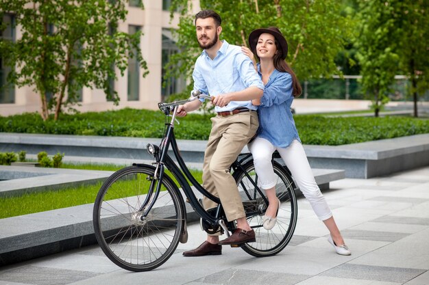 Pareja joven sentada en una bicicleta frente al parque verde de la ciudad