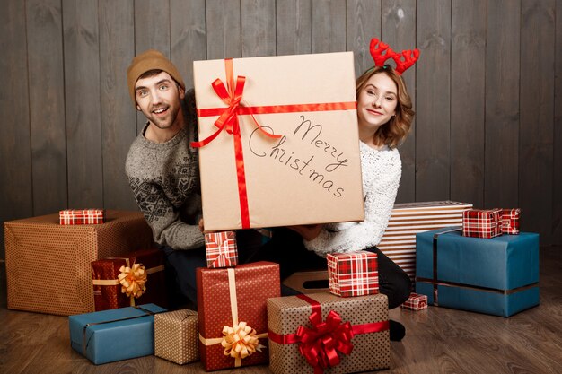 Pareja joven sentada entre cajas de regalo de Navidad sobre pared de madera