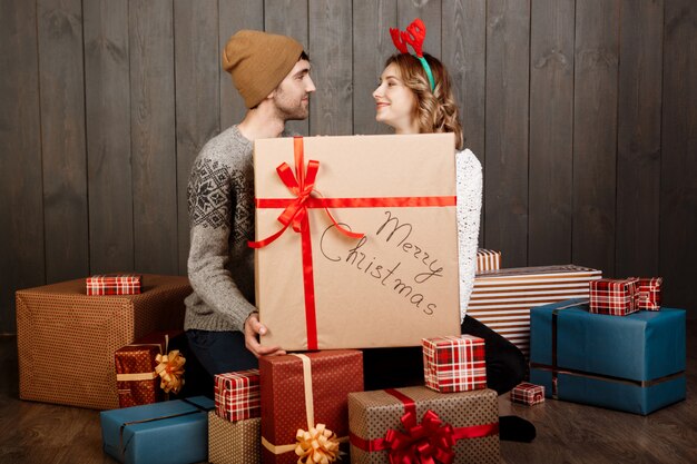 Pareja joven sentada entre cajas de regalo de Navidad sobre pared de madera