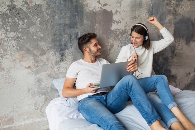 Pareja joven sentada en la cama en casa en ropa casual, hombre ocupado trabajando como autónomo en la computadora portátil, mujer escuchando música en auriculares, pasando tiempo juntos