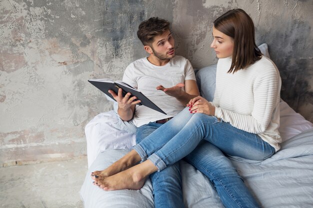 Pareja joven sentada en la cama en casa en ropa casual libro de lectura vistiendo jeans, discutiendo, problemas familiares