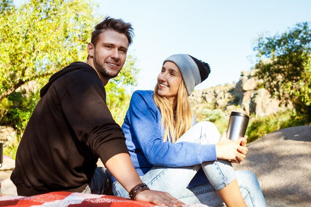 Pareja joven sentada en la roca en el cañón, sonriendo, bebiendo té