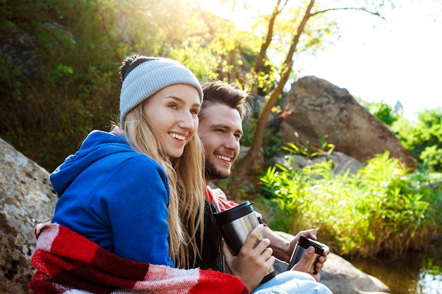 Pareja joven sentada en la roca en el cañón, sonriendo, bebiendo té