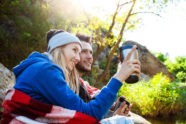 Pareja joven sentada en la roca en el cañón, sonriendo, bebiendo té