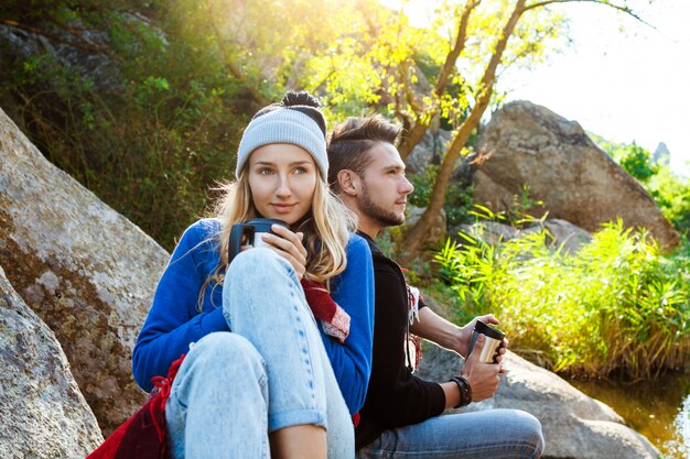 Pareja joven sentada en la roca en el cañón, sonriendo, bebiendo té