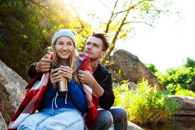 Pareja joven sentada en la roca en el cañón, sonriendo, bebiendo té