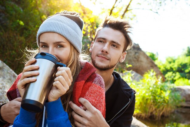 Pareja joven sentada en la roca en el cañón, sonriendo, bebiendo té