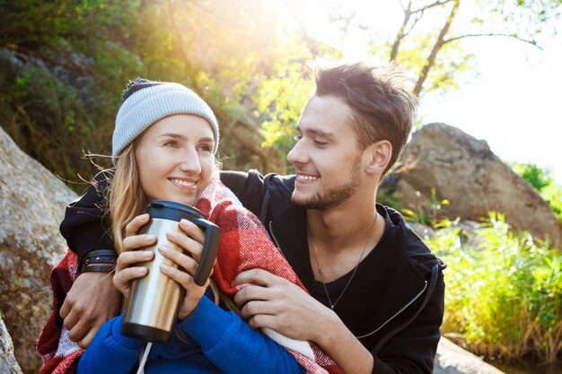 Pareja joven sentada en la roca en el cañón, sonriendo, bebiendo té