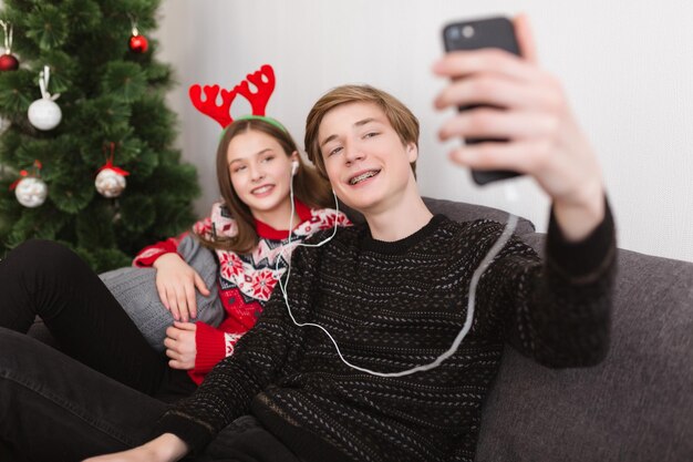 Pareja joven sentada en un sofá en casa con auriculares y tomando selfie mientras pasa tiempo junto con el árbol de Navidad en el fondo