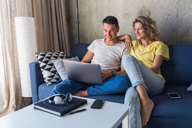 Pareja joven sentada en el sofá en casa mirando en la computadora portátil, viendo películas en línea, usando internet