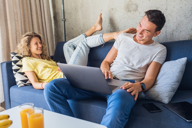 Pareja joven sentada en el sofá en casa usando laptop, jugando y coqueteando