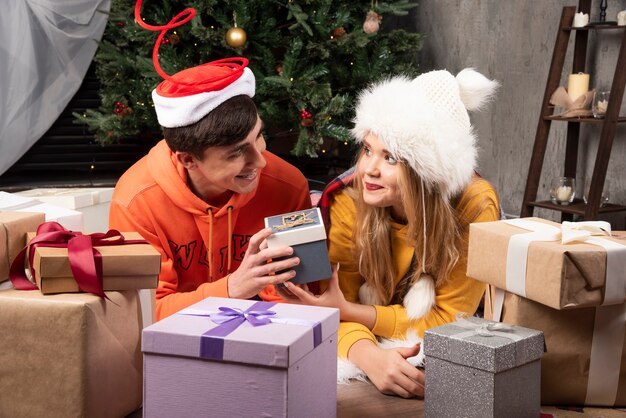 Pareja joven sentada en el suelo y posando con regalos cerca del árbol de Navidad