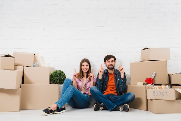 Pareja joven sentada en el suelo sosteniendo sus dedos hacia arriba, sentada entre las cajas de cartón