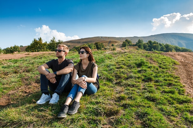 Pareja joven sonriendo, disfrutando del paisaje de montañas, sentado en la colina