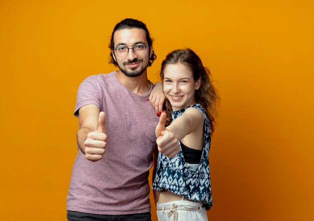 Pareja joven sonriendo feliz y positivo mostrando Thumbs up parado sobre pared naranja