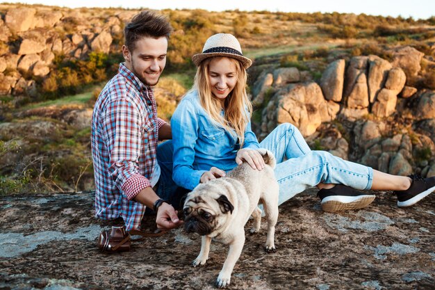 Pareja joven sonriendo, sentado en la roca en el cañón, acariciando perro pug