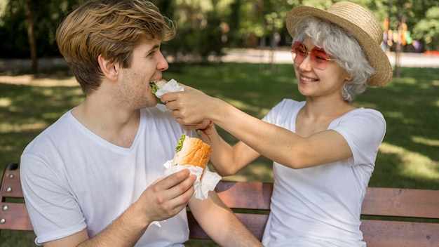 Pareja joven sonriente comiendo hamburguesas en el parque