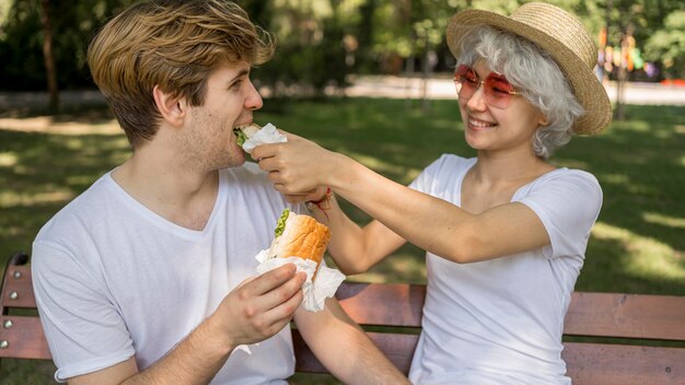 Pareja joven sonriente comiendo hamburguesas en el parque