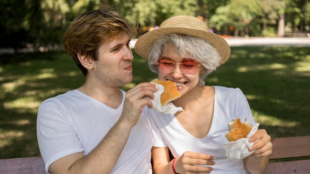 Pareja joven sonriente comiendo hamburguesas en el parque