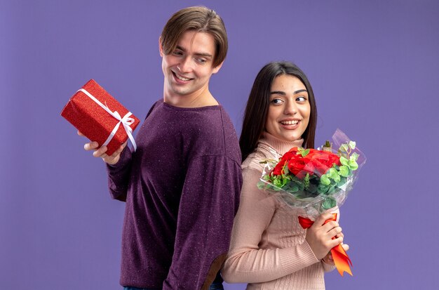 Pareja joven sonriente en el día de San Valentín reteniendo espalda con espalda con caja de regalo y ramo aislado sobre fondo azul.