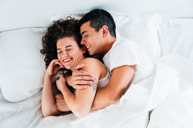 Pareja joven sonriente bajo edredón en cama