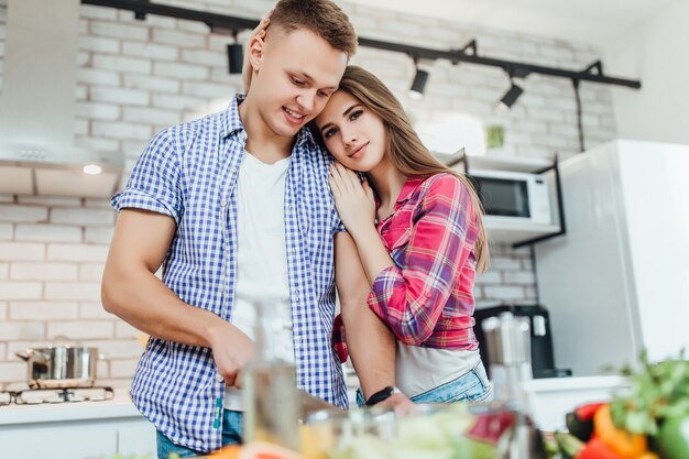 Pareja joven sonriente preparando la cena. El hombre está cortando verduras con un cuchillo, la mujer abraza la suya por detrás.