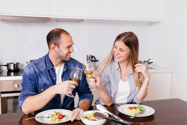 Pareja joven sonriente que come la ensalada que tuesta con las copas de vino en la cocina