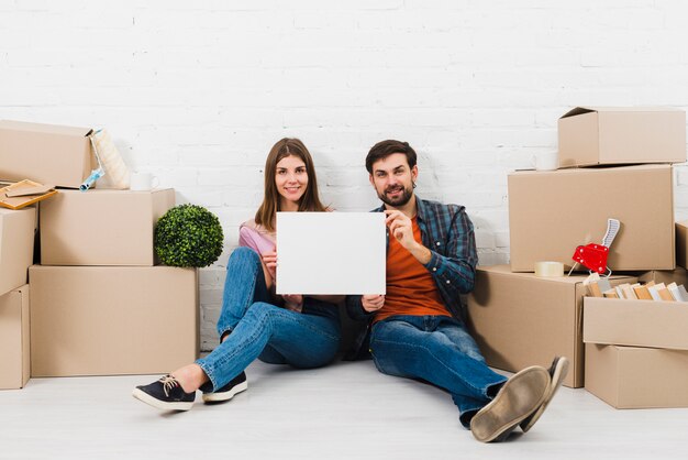 Pareja joven sonriente que muestra el cartel blanco en blanco que se sienta entre las cajas de cartón